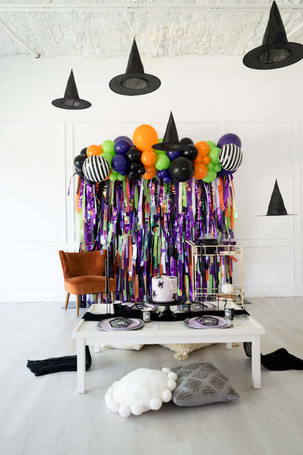 Halloween party decor featuring vibrant fringe backdrops, balloons, black hats, and a spooky cake.