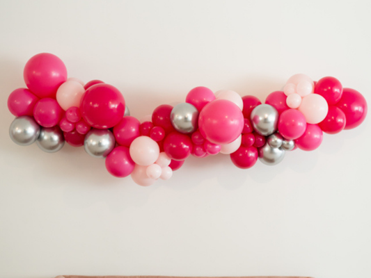 Pink Doll Balloon Kit fringe garland hanging against a white wall with balloons. Fringe decor perfect for corporate events or parties.