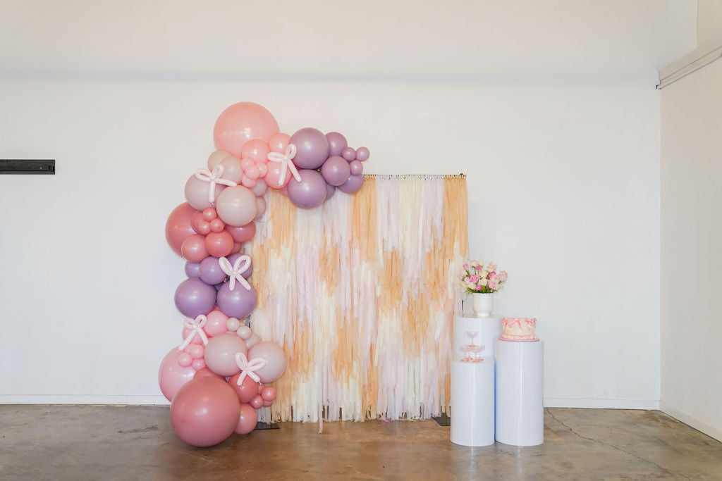 Bashful Blossoms Backdrop fringe garland hanging against a white wall with balloons. Fringe decor perfect for corporate events or parties.